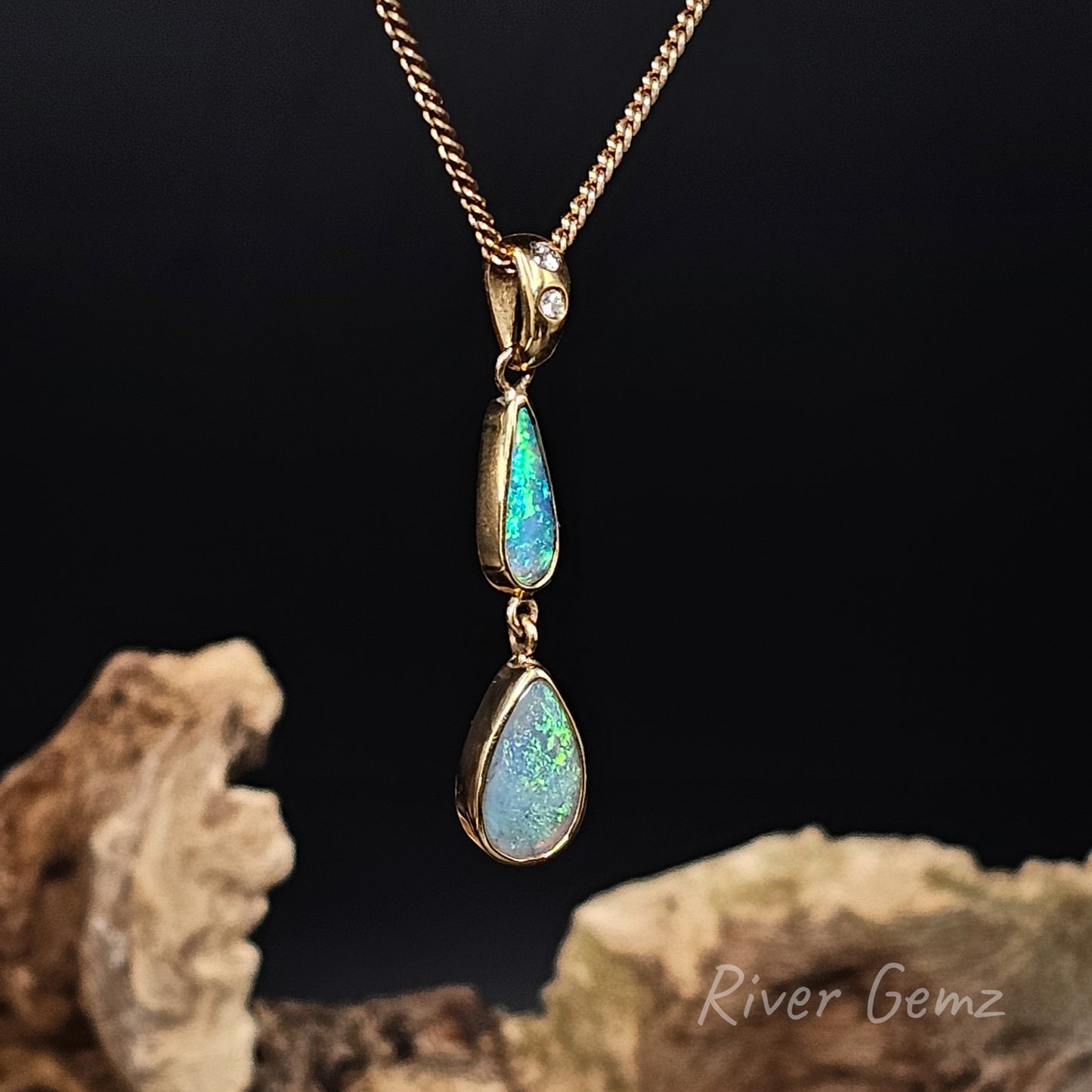 A gold pendant necklace featuring two dangling teardrop-shaped iridescent opal gemstones, displayed against a dark background with decorative driftwood and the text 'River Gemz'.
