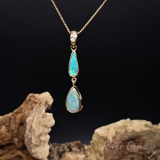 A gold pendant necklace featuring two teardrop-shaped iridescent opal gemstones, displayed on a dark background with decorative driftwood. The brand name 'River Gemz' is visible in the bottom right corner.