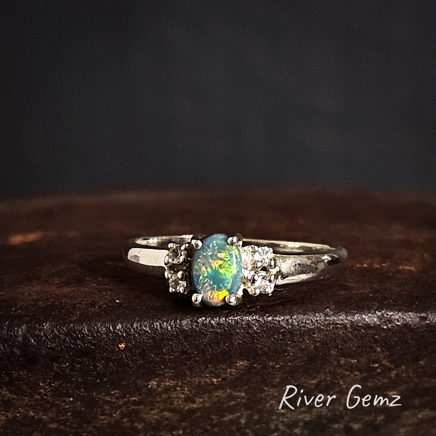 Silver ring with a multi-coloured black opal and white topaz stones on a dark rusty surface, branded 'River Gemz'.