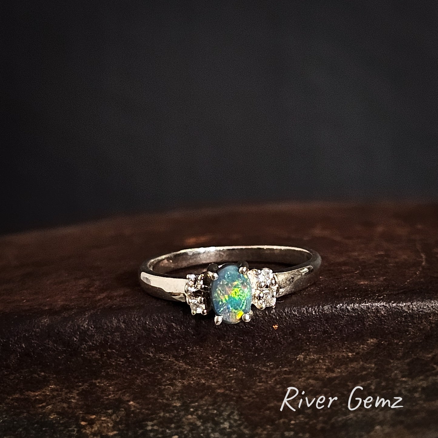 Silver ring with a green, yellow and blue dark opal on a dark brown surface, branded 'River Gemz'.