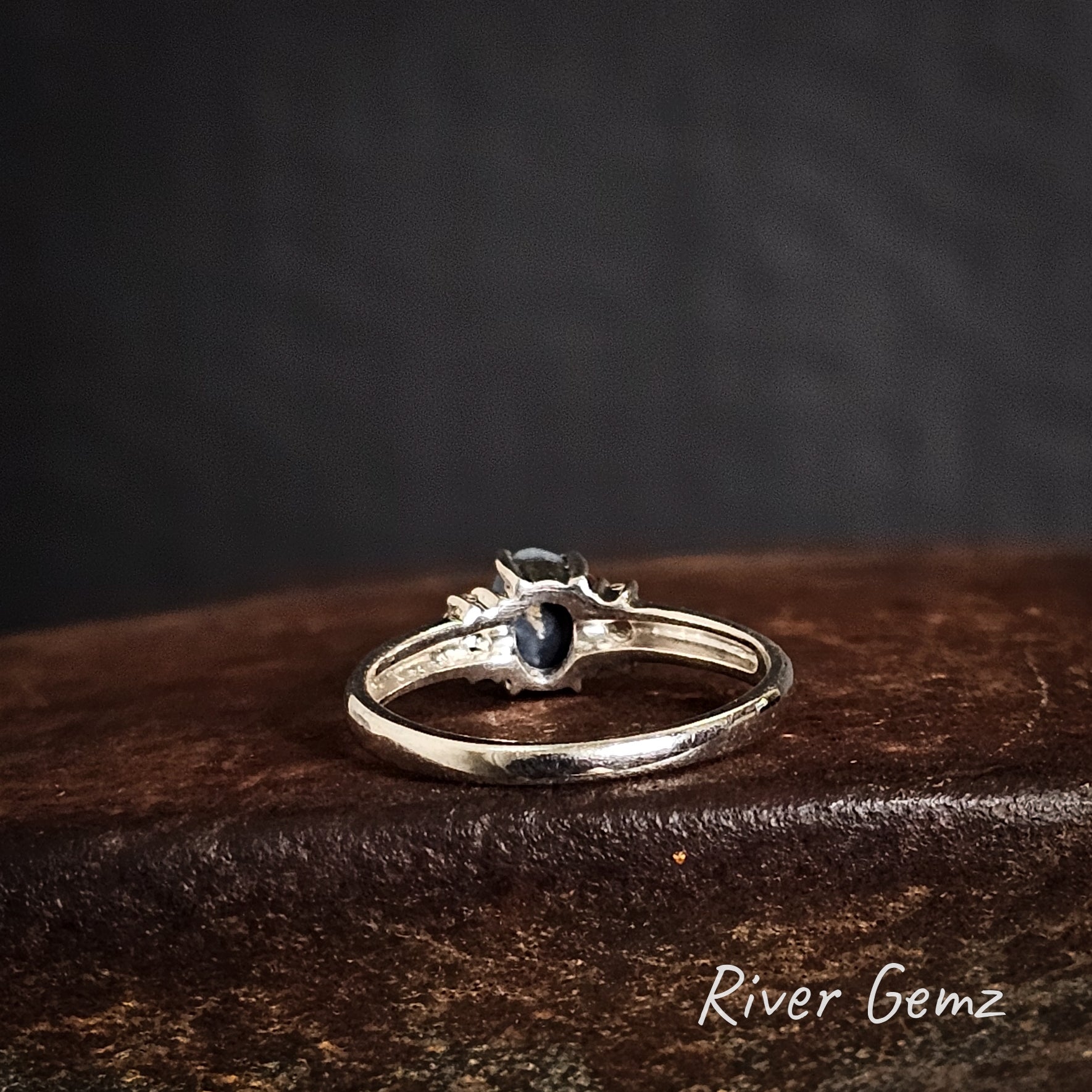 Rear view of silver ring with a black gemstone on a dark brown surface, branded 'River Gemz'.