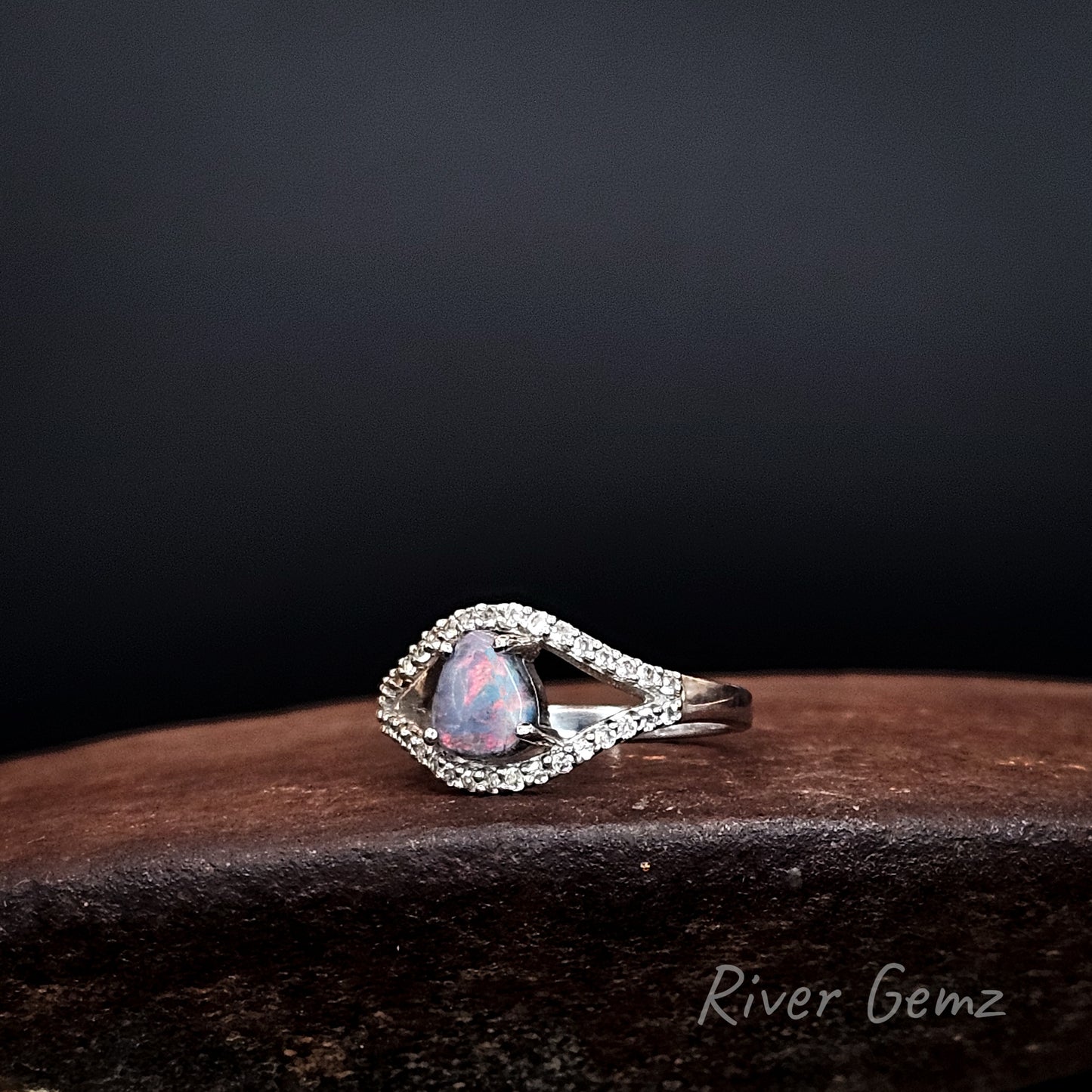 Rounded corner triangular shaped opal with dominant reds claw set in sterling silver ring with a halo of white topaz stones.