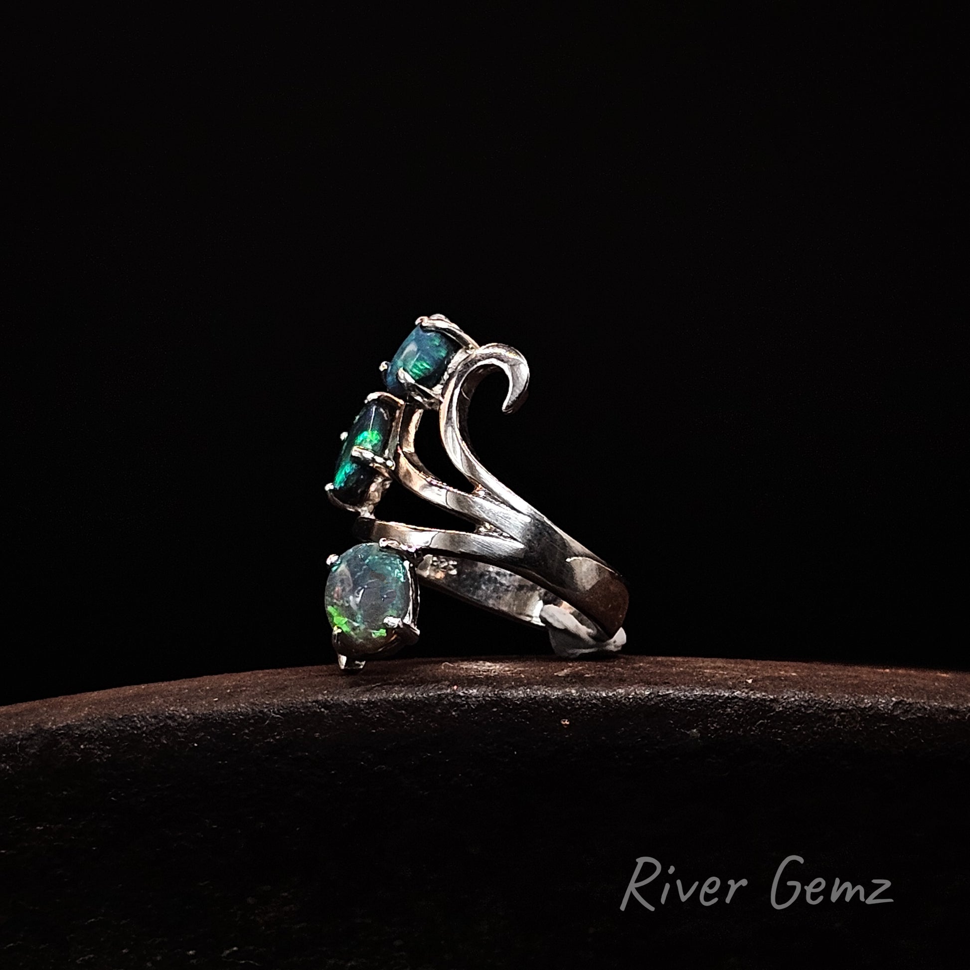 A silver ring with three cascading green opal gemstones, displayed on a dark brown surface against a solid black background. The brand name 'River Gemz' is visible in the bottom right corner.