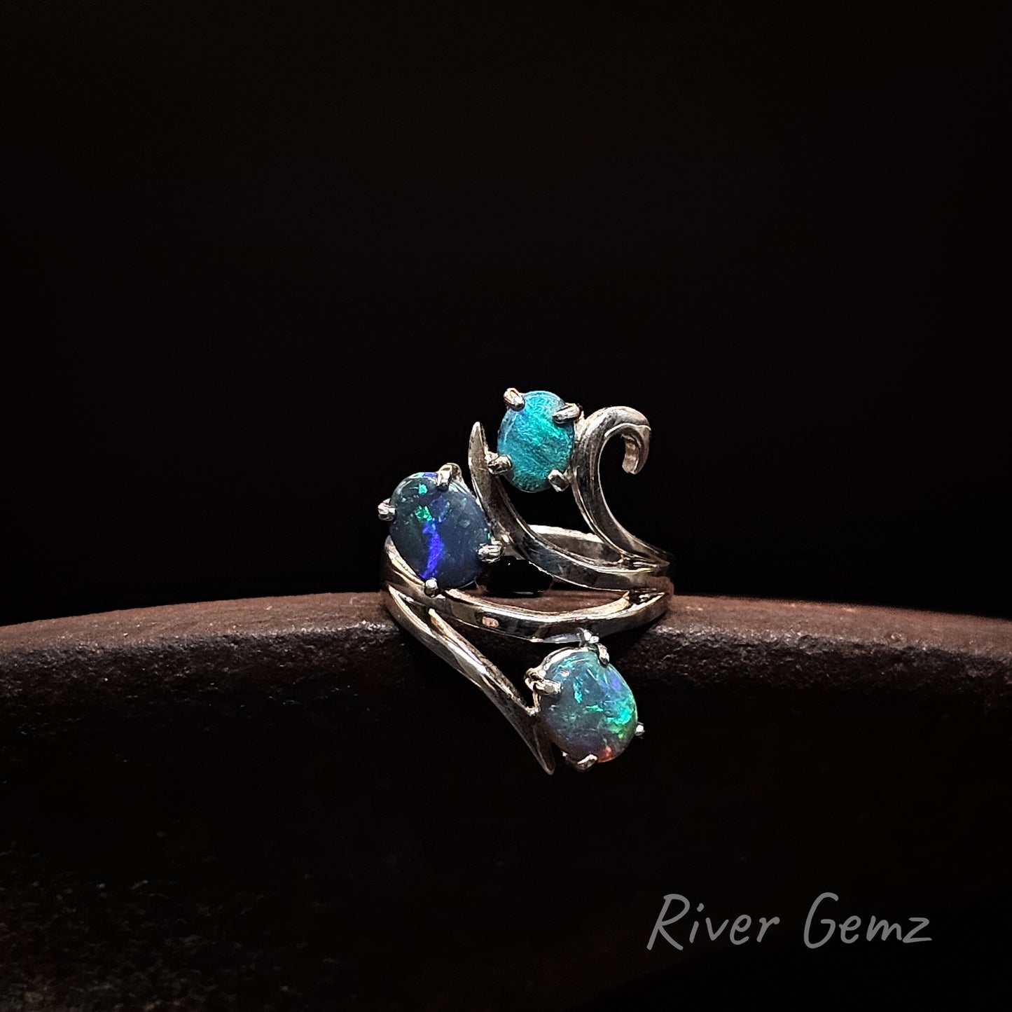 A silver ring with three iridescent blue-green opal gemstones, displayed on a dark brown textured surface against a black background. The brand name 'River Gemz' is visible in the bottom right corner.
