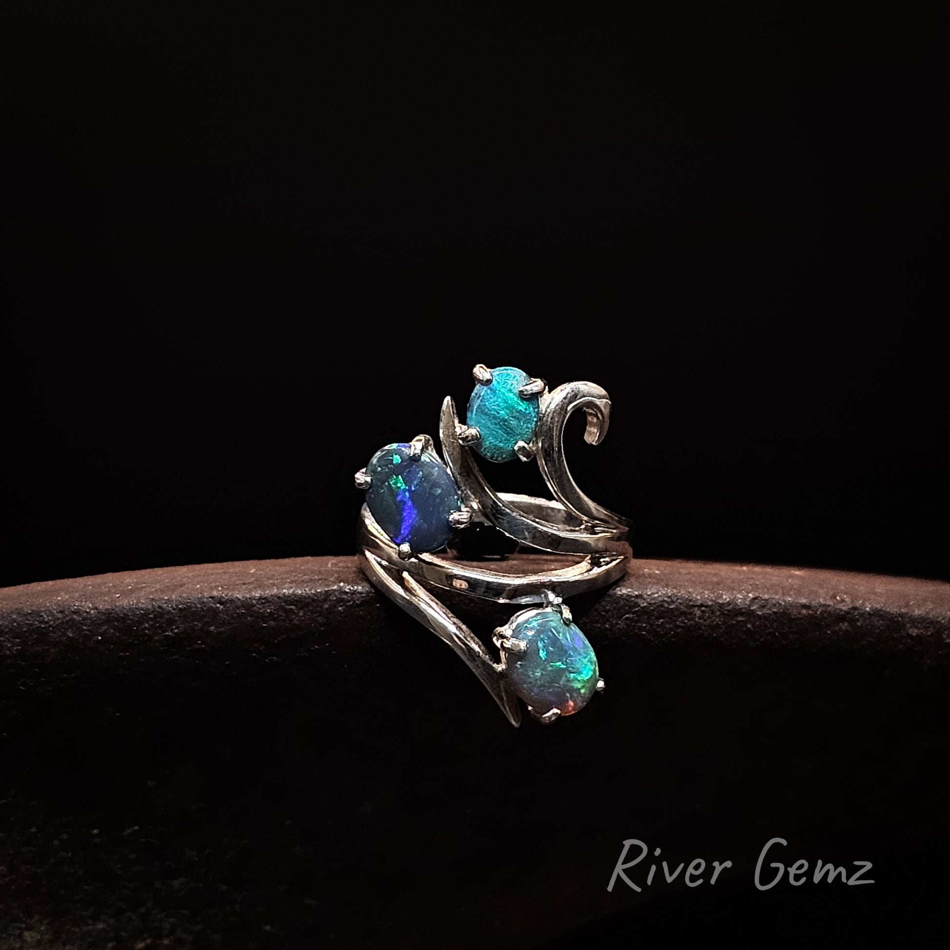 A silver ring with three iridescent blue-green opal gemstones, displayed on a dark brown textured surface against a black background. The brand name 'River Gemz' is visible in the bottom right corner.