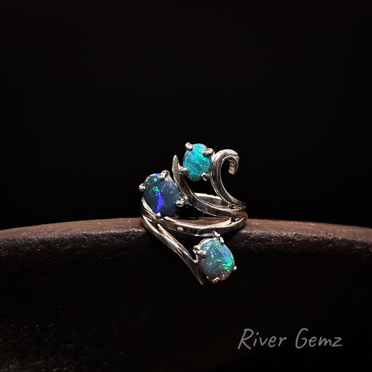 A silver ring with three iridescent blue-green opal gemstones, displayed on a dark brown textured surface against a black background. The brand name 'River Gemz' is visible in the bottom right corner.