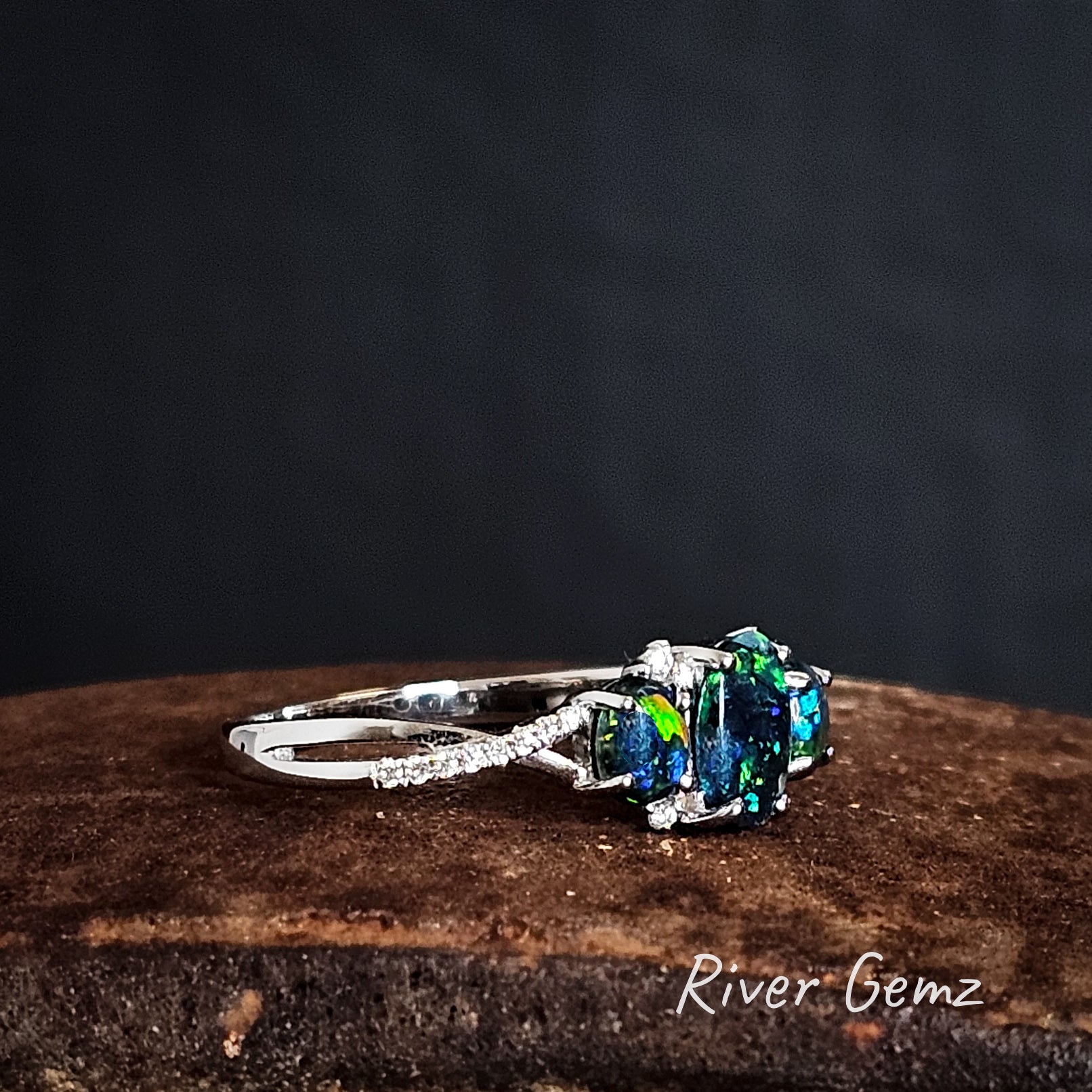 Ring with green and blue dark opal on a rust coloured surface, branded 'River Gemz'.