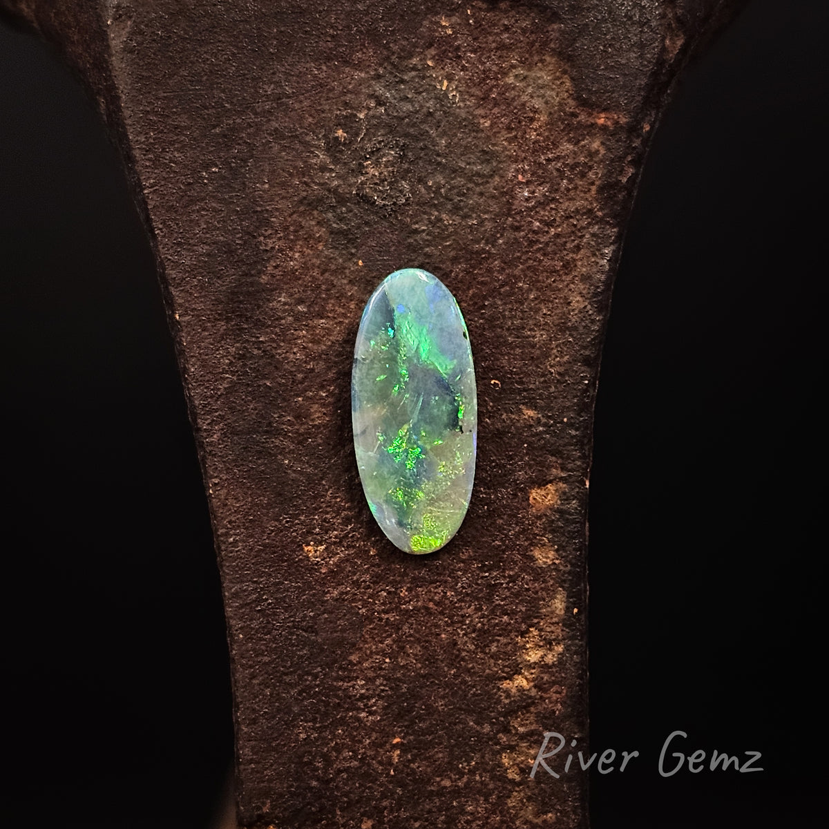 Oval shaped blue-green opal on a rusty shoe anvil with 'River Gemz' watermark in the bottom right hand corner against a black background.