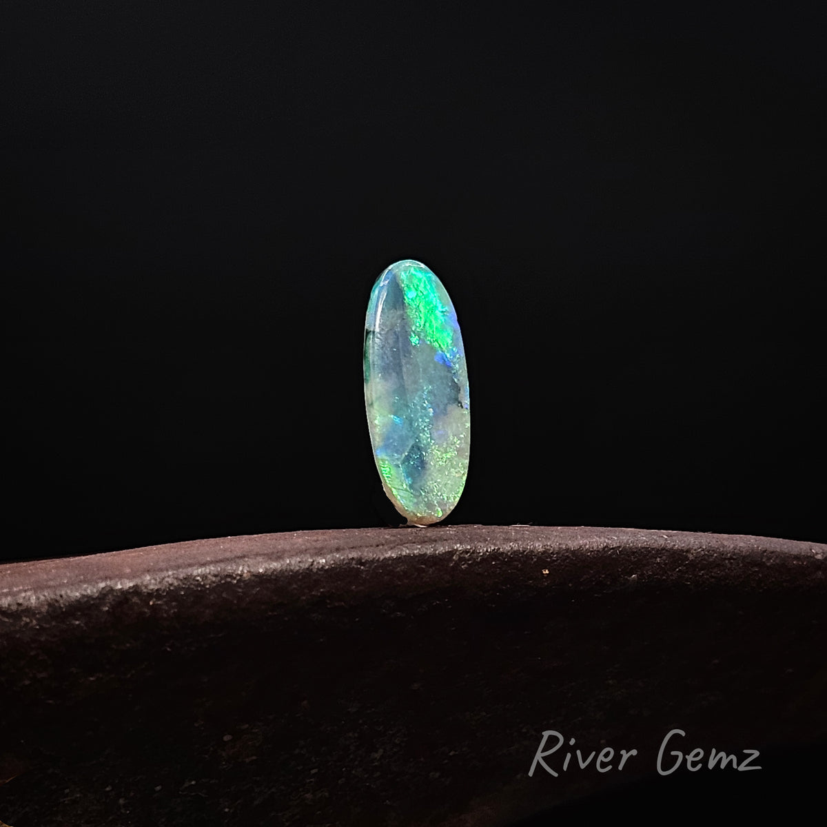 Oval shaped blue-green opal standing upright on a rusty shoe anvil with 'River Gemz' watermark in the bottom right hand corner against a black background.