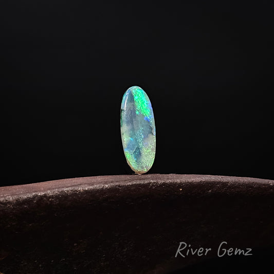 Oval shaped blue-green opal standing upright on a rusty shoe anvil with 'River Gemz' watermark in the bottom right hand corner against a black background.