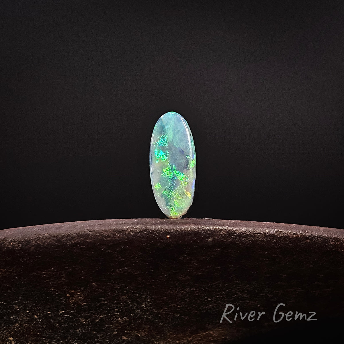 Oval shaped blue-green opal standing upright on a rusty shoe anvil with 'River Gemz' watermark in the bottom right hand corner against a black background.