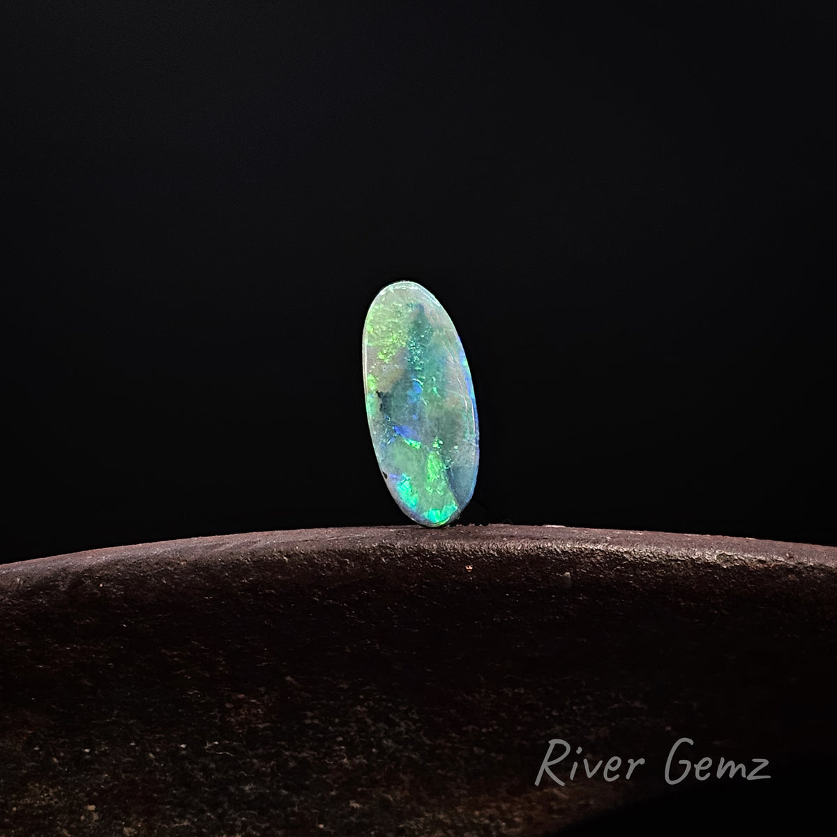 Oval shaped blue-green opal standing upright on a rusty shoe anvil with 'River Gemz' watermark in the bottom right hand corner against a black background.