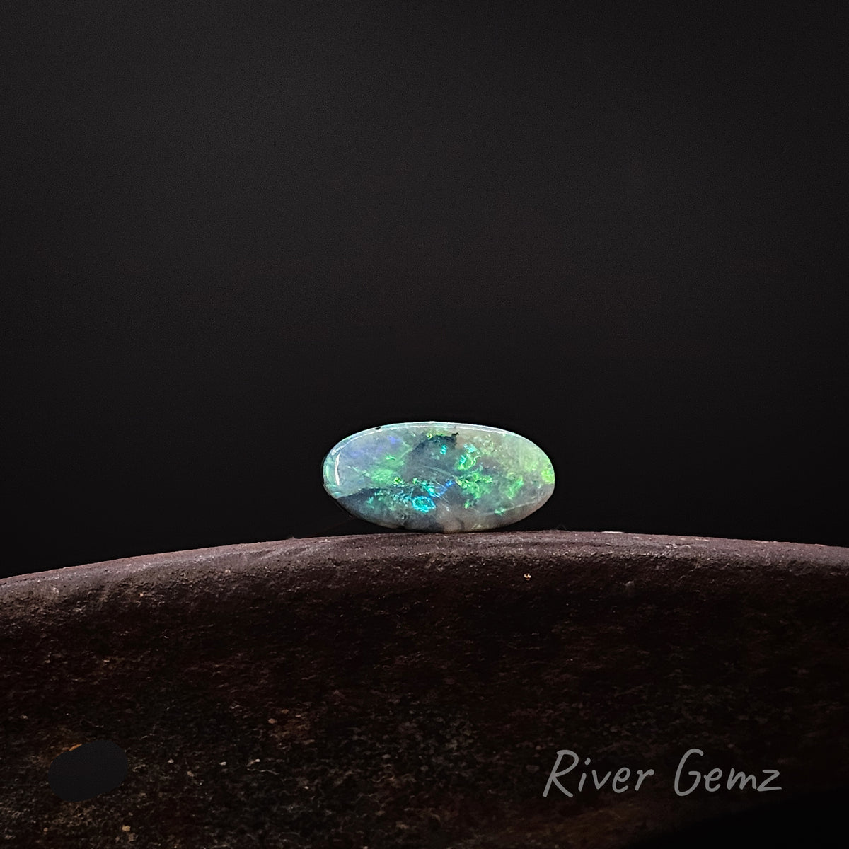 Oval shaped blue-green opal shown horizontally [East to West] on a rusty shoe anvil with 'River Gemz' watermark in the bottom right hand corner against a black background.