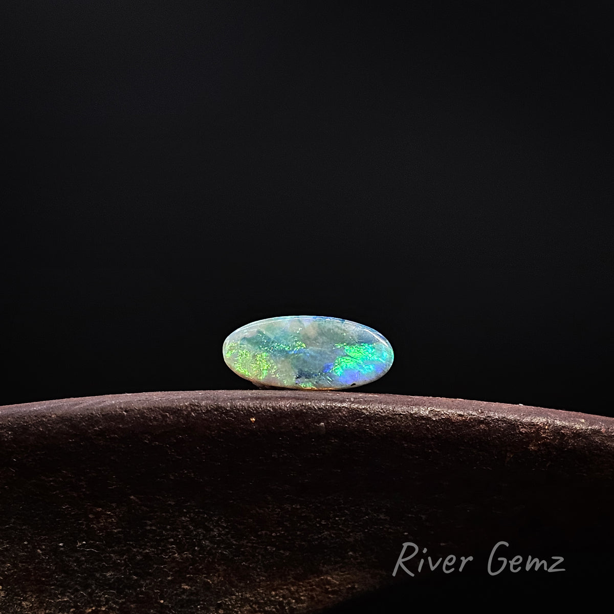Oval shaped blue-green opal shown horizontally [East to West] on a rusty shoe anvil with 'River Gemz' watermark in the bottom right hand corner against a black background.