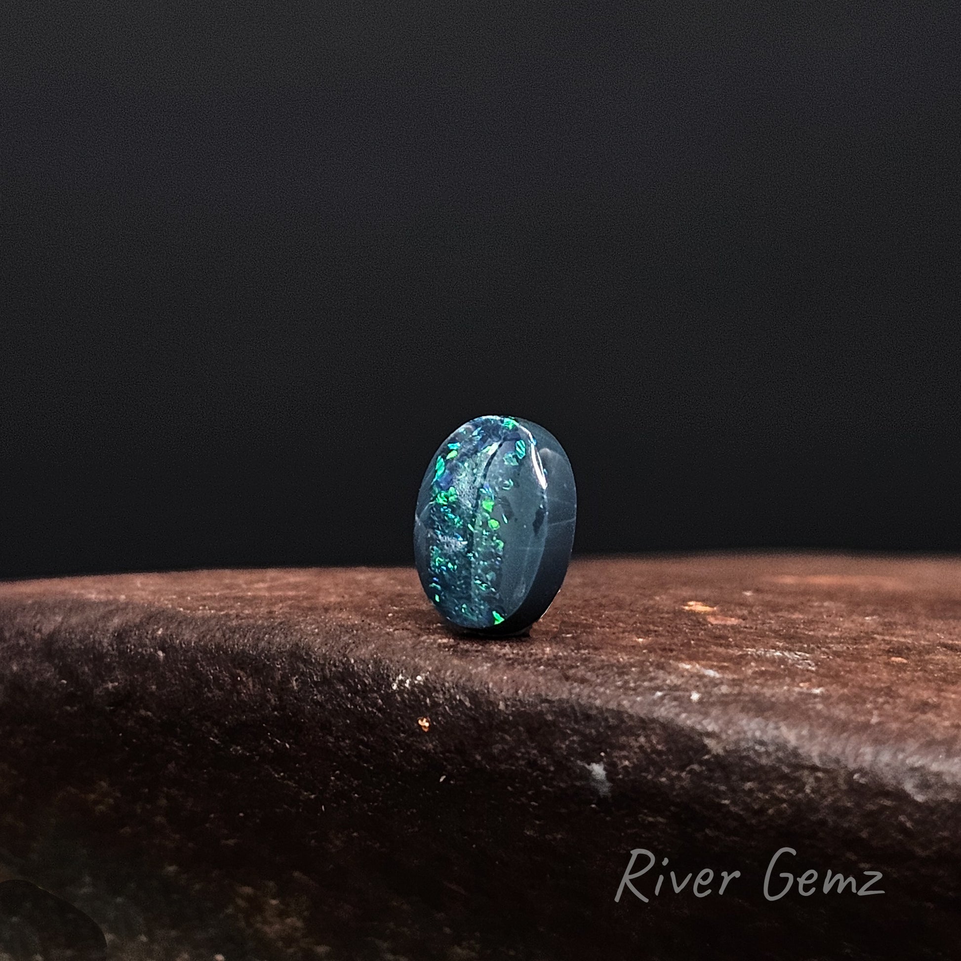 Loose opal stone on a dark surface with a black background, branded 'River Gemz'.