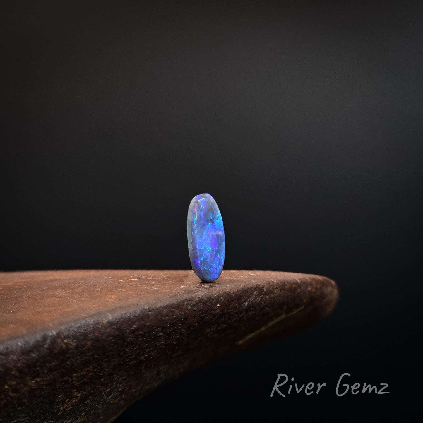Oval shaped blue, purple and green black opal upright on a shoe anvil and black background with 'River Gemz' branding.
