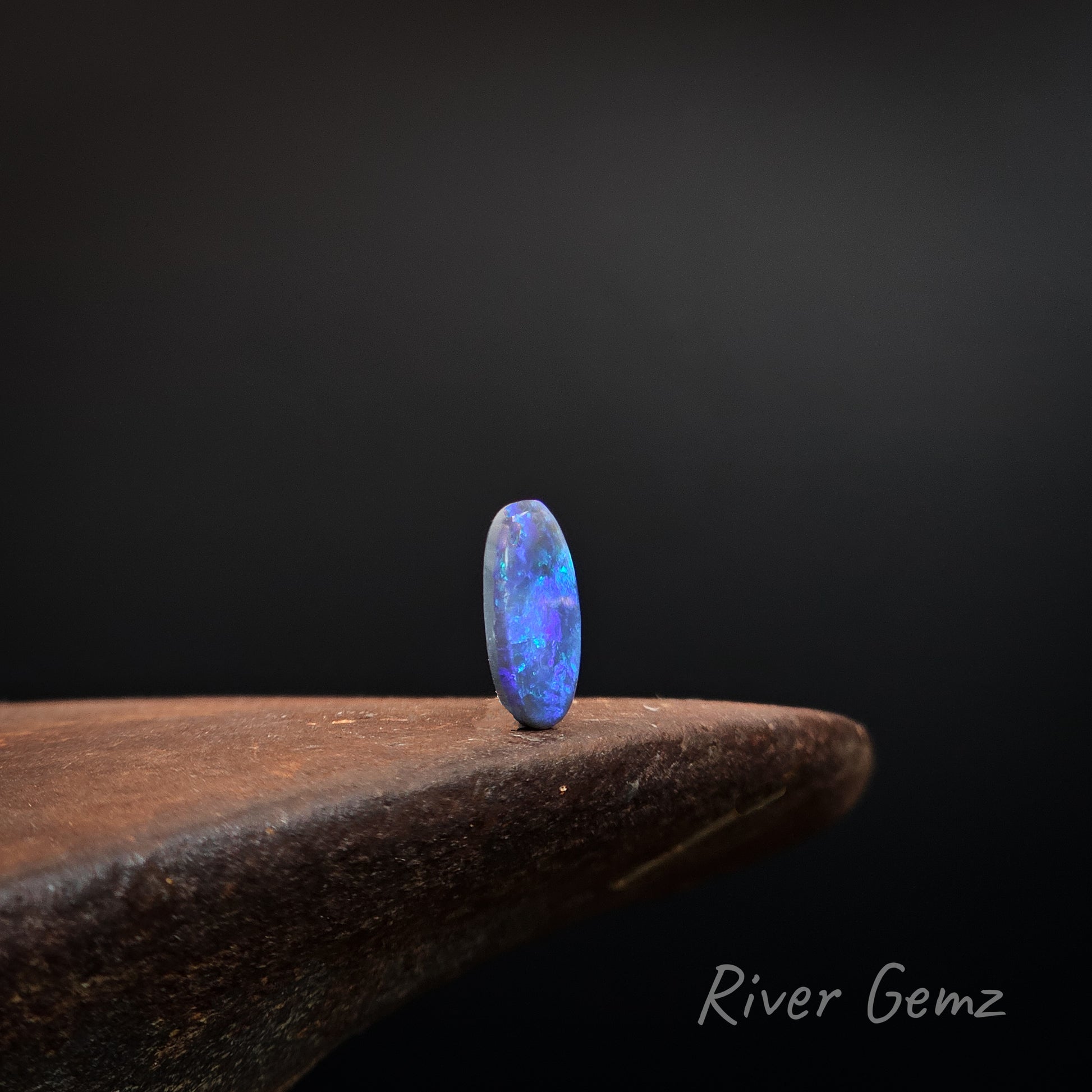 Oval shaped blue, purple and green black opal upright on a shoe anvil and black background with 'River Gemz' branding.