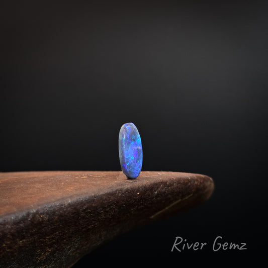 Oval shaped blue, purple and green black opal upright on a shoe anvil and black background with 'River Gemz' branding.