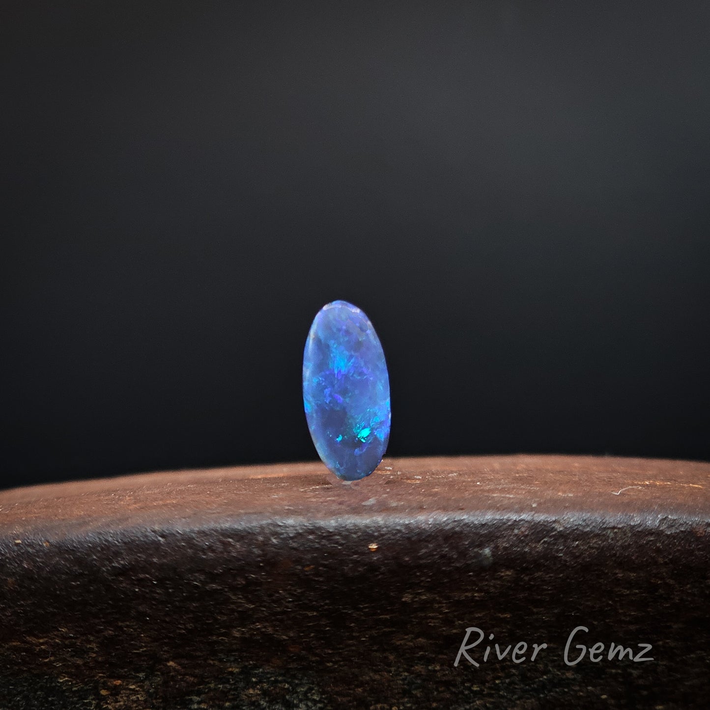 Predominantly blue coloured unset black opal standing upright on a brown surface with 'River Gemz' watermark.