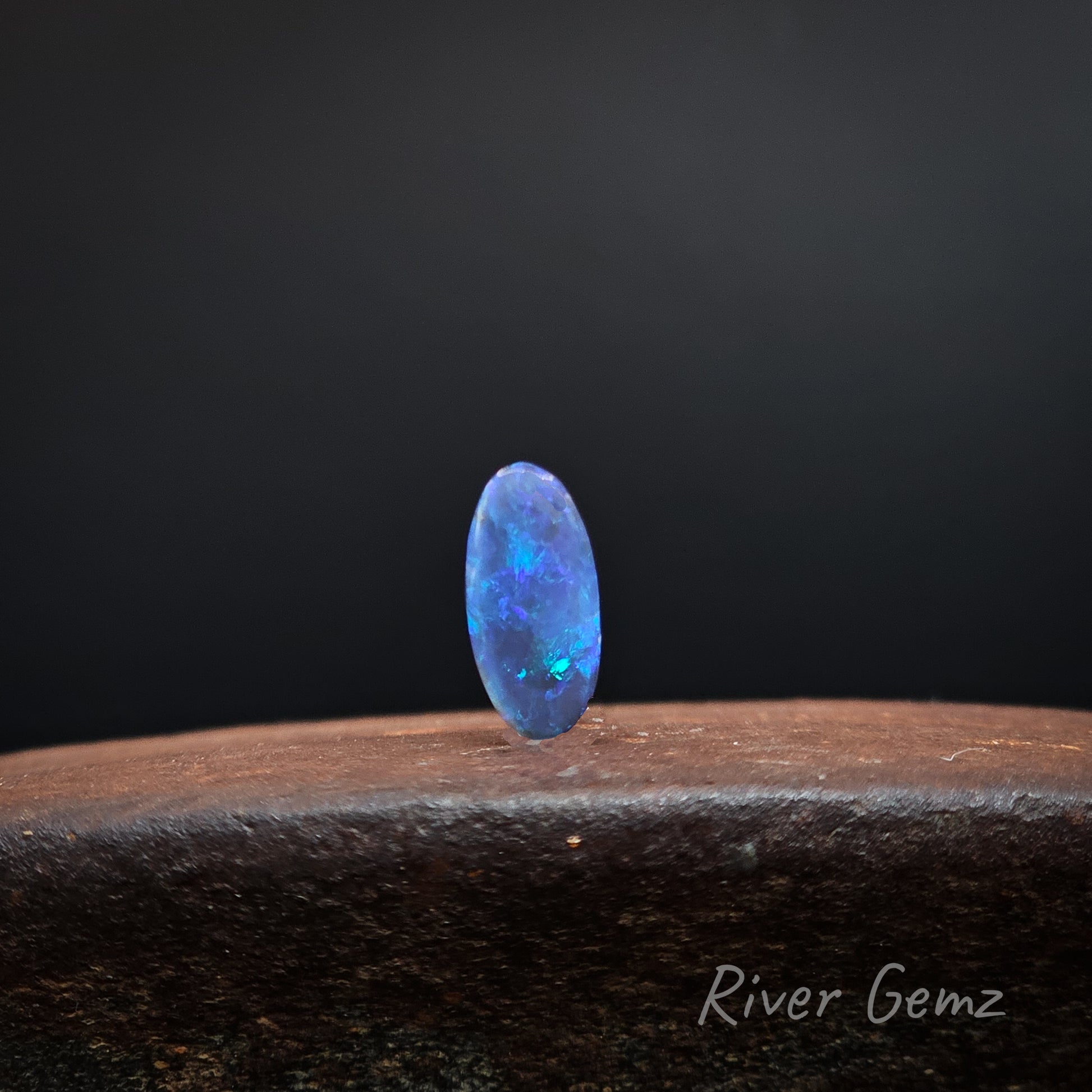 Predominantly blue coloured unset black opal standing upright on a brown surface with 'River Gemz' watermark.