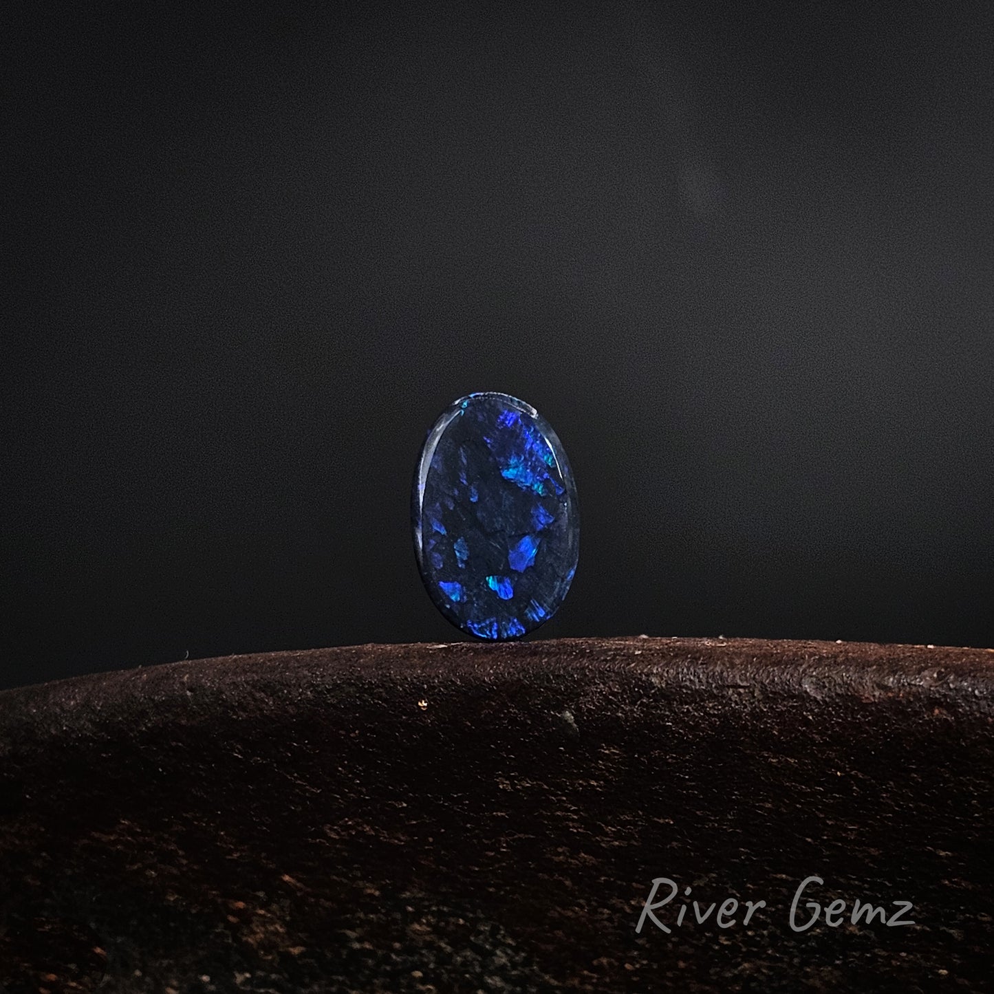 Oval shaped blue and green black opal on a brown base and dark grey background.
