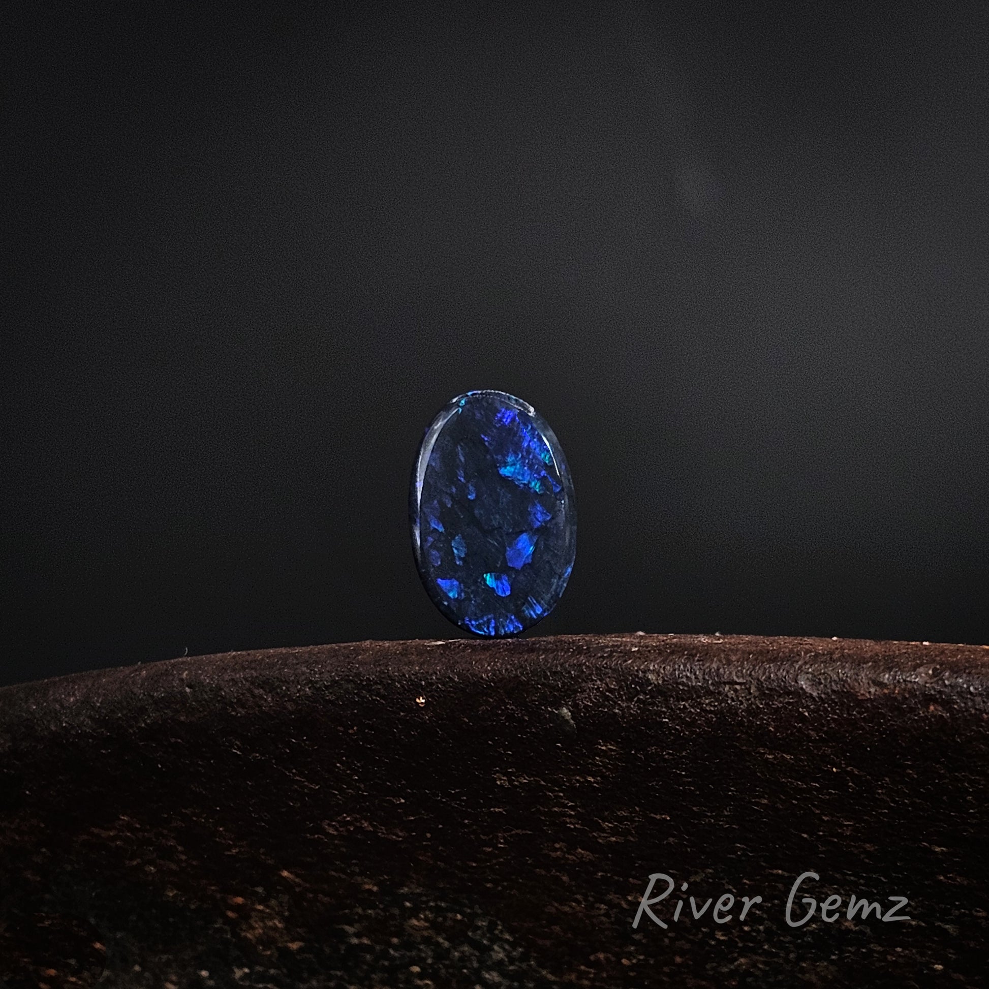 Oval shaped blue and green black opal on a brown base and dark grey background.