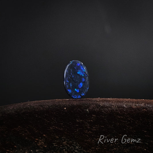 Oval shaped blue and green black opal on a brown base and dark grey background.