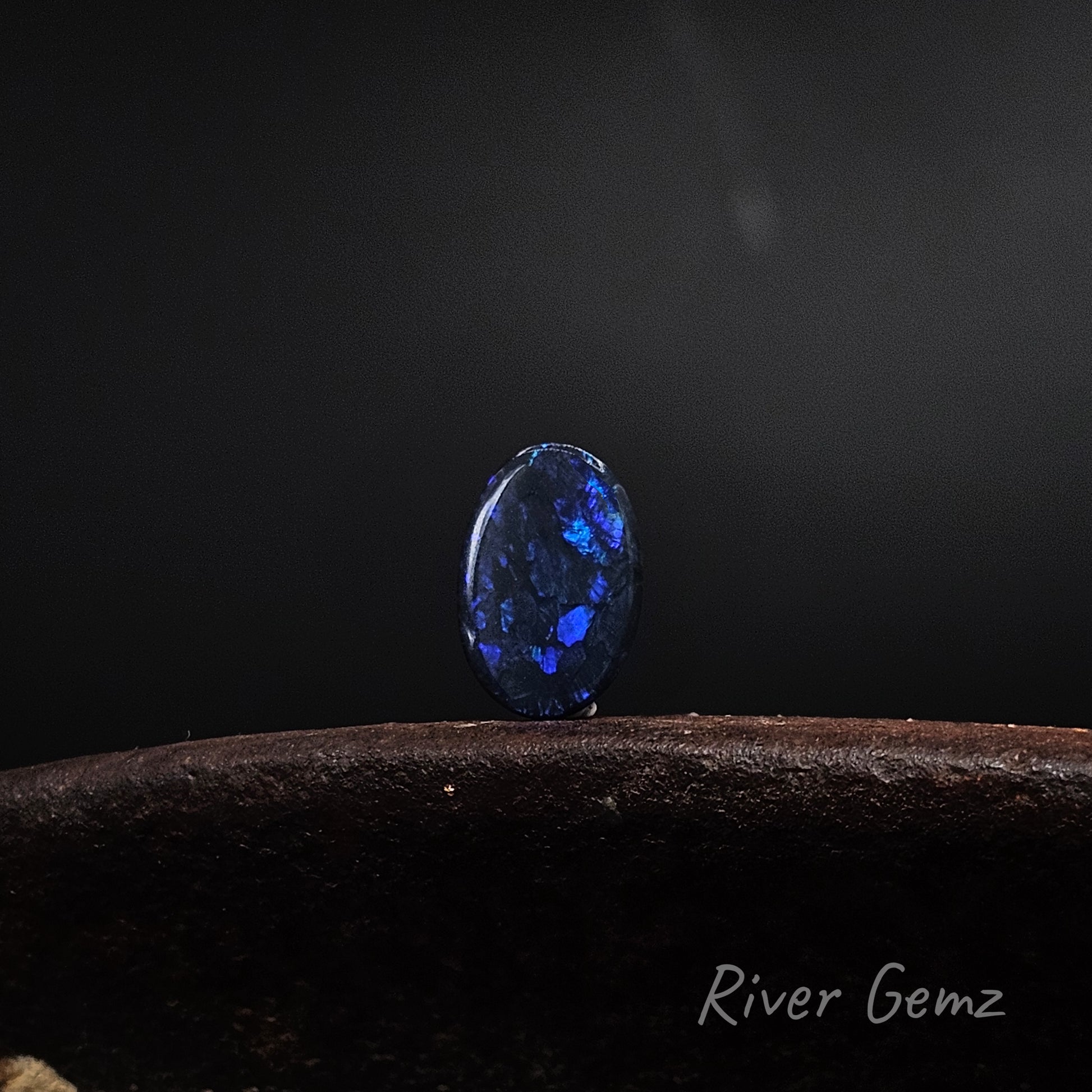 Dark blue spheres of colour in oval shaped loose opal standing upright on rusty shoe anvil with 'River Gemz' watermark in bottom right hand corner.