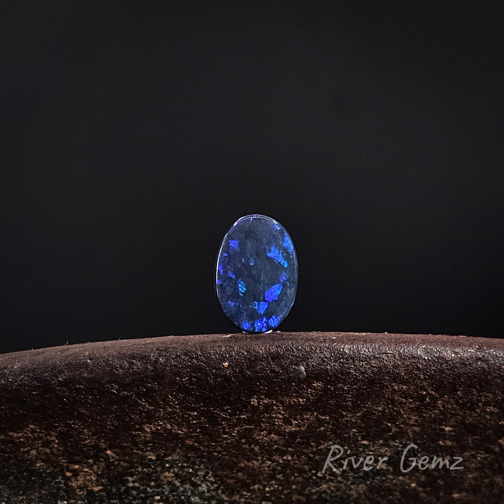 Unset dark opal with large spheres of dark blue, green and purple colours. The stone is standing upright on a shoe anvil with 'River Gemz' branding in the bottom right hand corner.