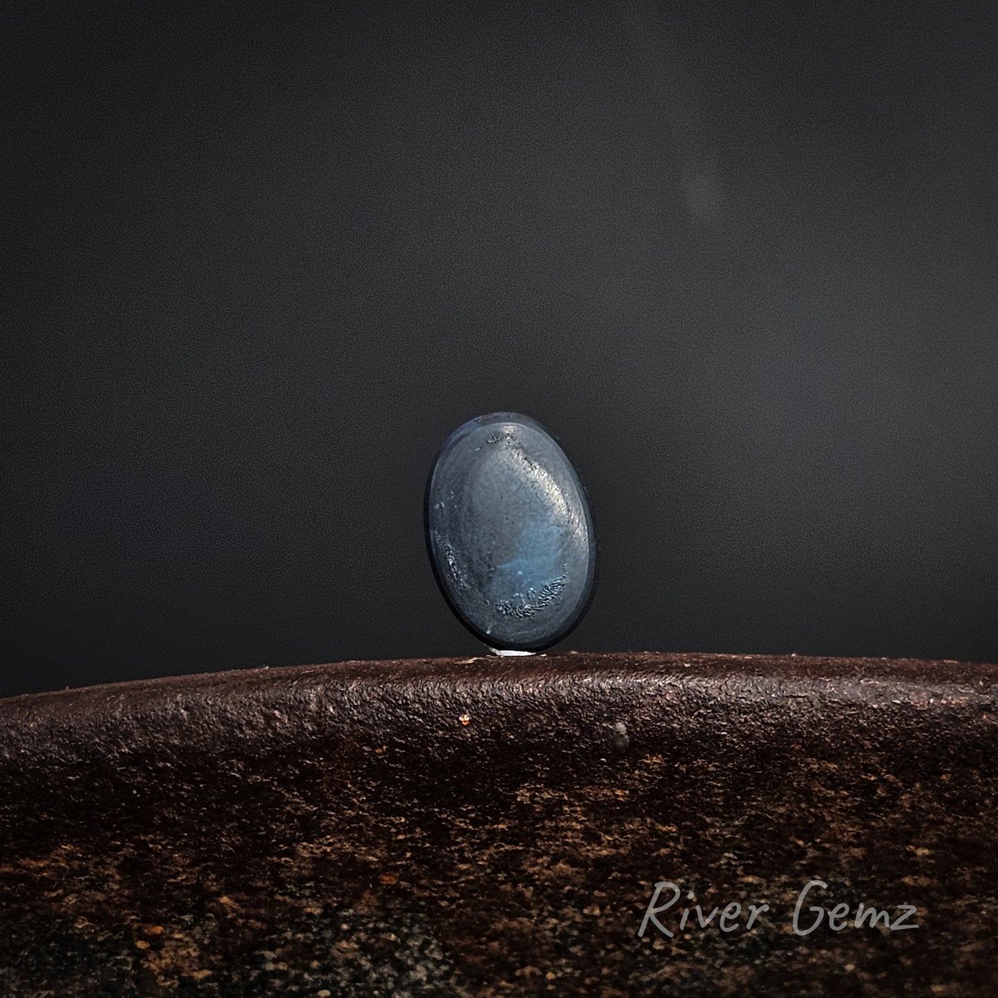 Rear view of the dark opal showing the dark base tone of the unset black opal standing upright on a brown base and dark grey background.