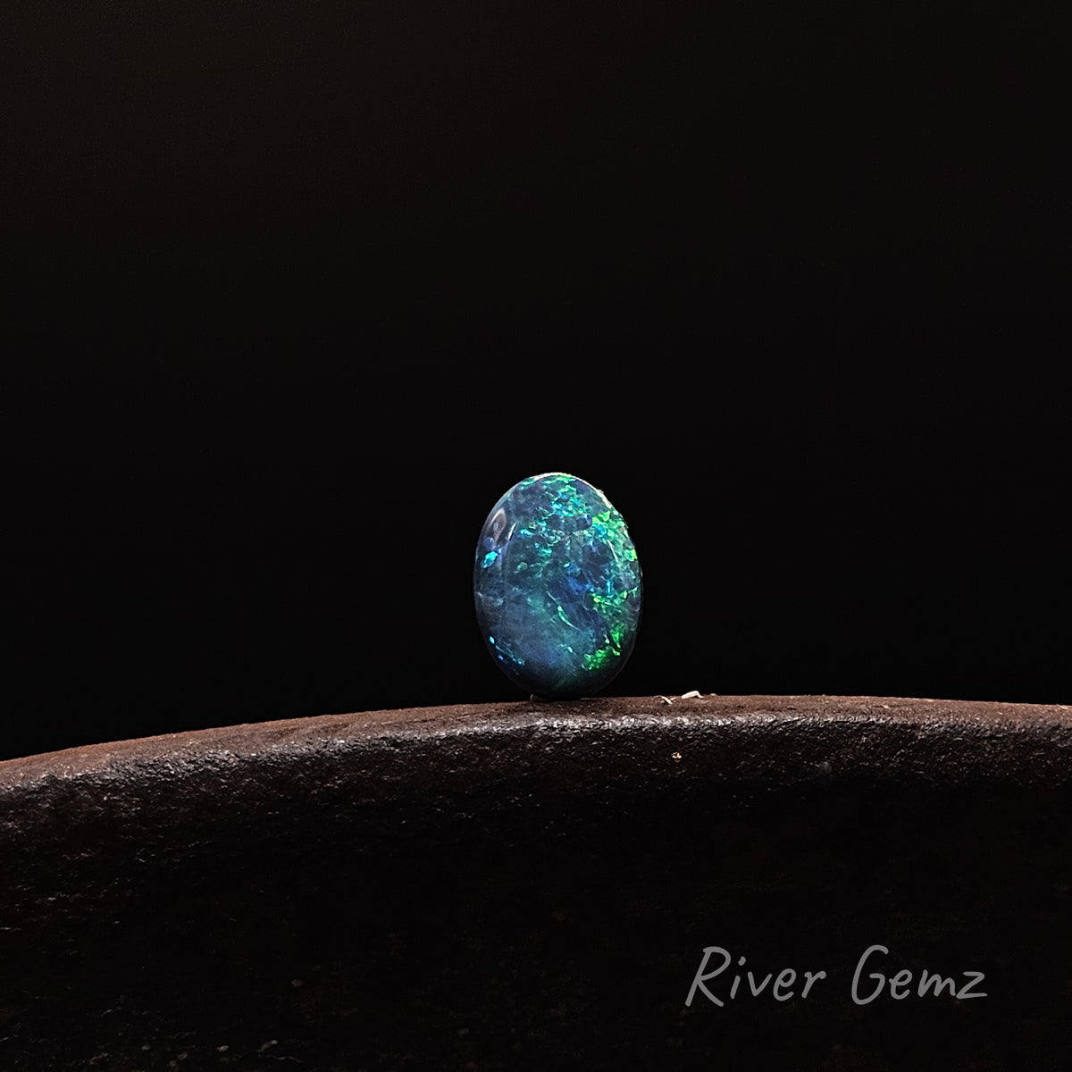 A single oval cabochon gemstone with vibrant blue, green, and black opal-like play-of-color, resting on a dark textured surface against a solid black background. The watermark 'River Gemz' is visible in the bottom right corner.
