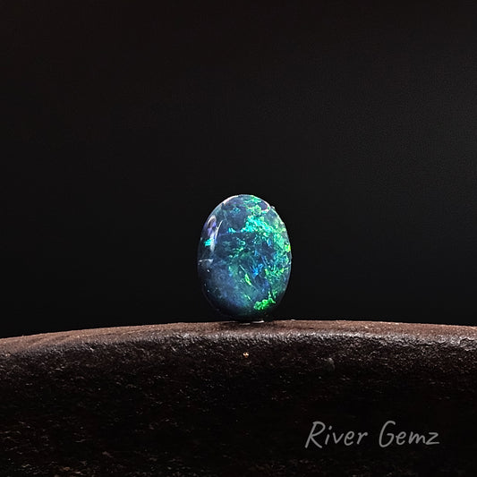 A polished oval-cut blue-green opal gemstone with vibrant iridescent flashes, displayed on a dark textured surface against a solid black background. The brand name 'River Gemz' is visible in the bottom right corner.