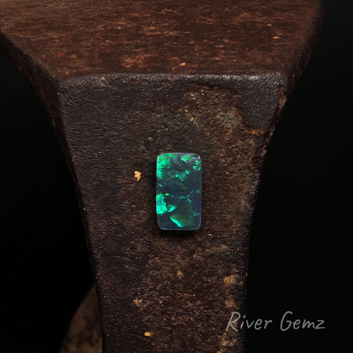 Rectangular shaped blue and green black opal against a rust coloured shoe anvil with 'River Gemz' watermark in the bottom right hand corner.