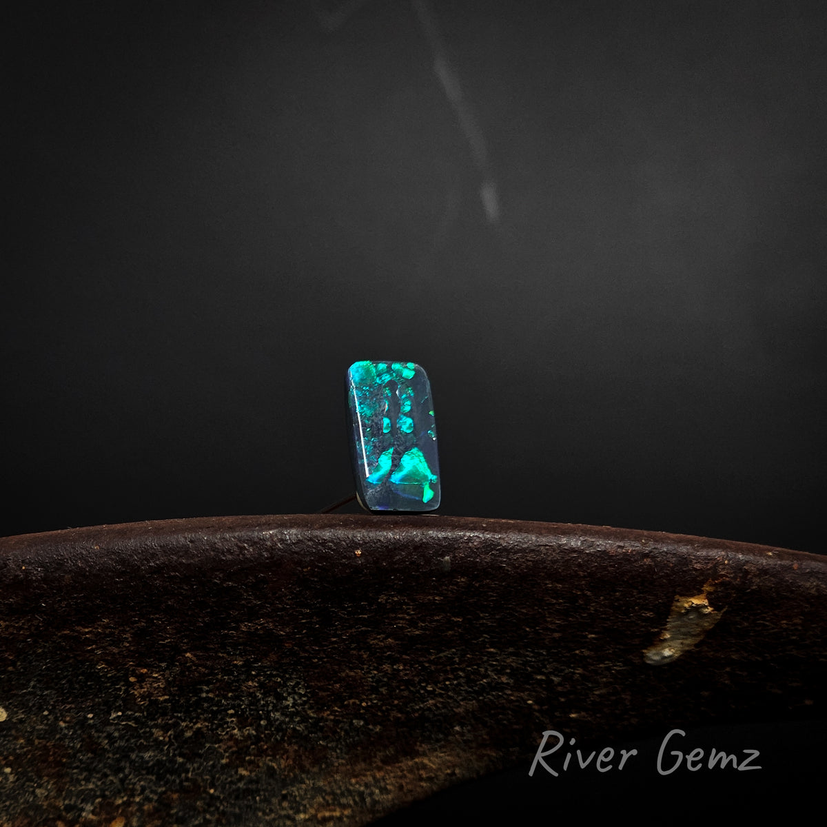 Rectangular shaped blue and green black opal standing upright on a brown base with 'River Gemz' watermark against a dark background.