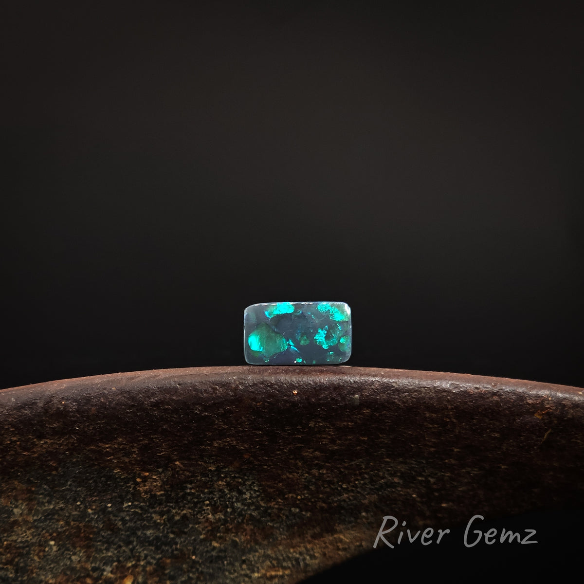 Rectangular shaped blue and green black opal shown in an East-West direction on a brown base with 'River Gemz' watermark against a dark background.