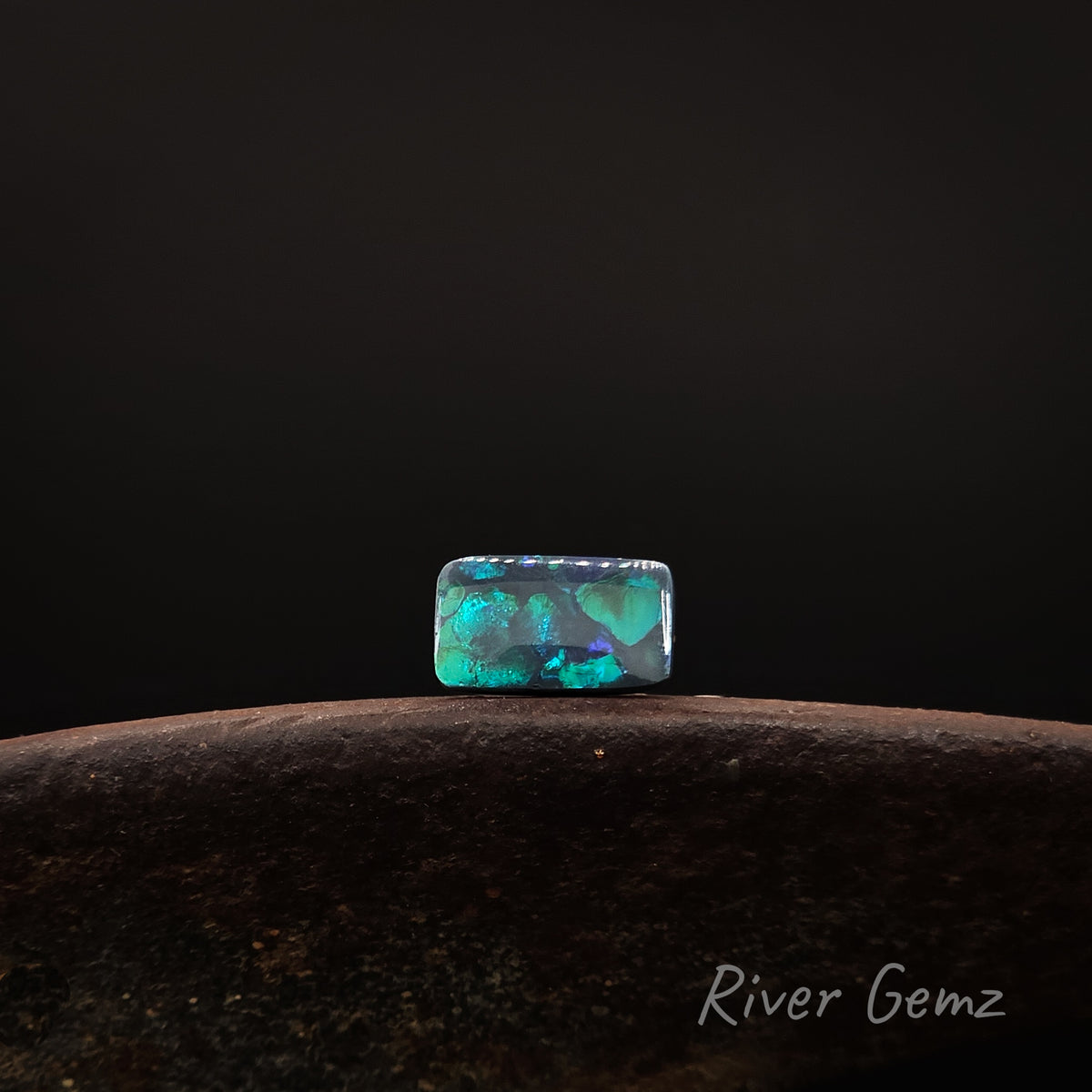 Rectangular shaped blue and green black opal shown in an East-West direction on a brown base with 'River Gemz' watermark against a dark background.