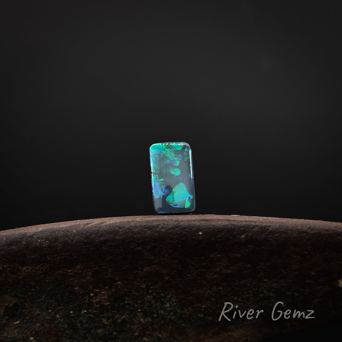 Rectangular shaped blue and green black opal standing upright on a brown base with 'River Gemz' watermark against a dark background.