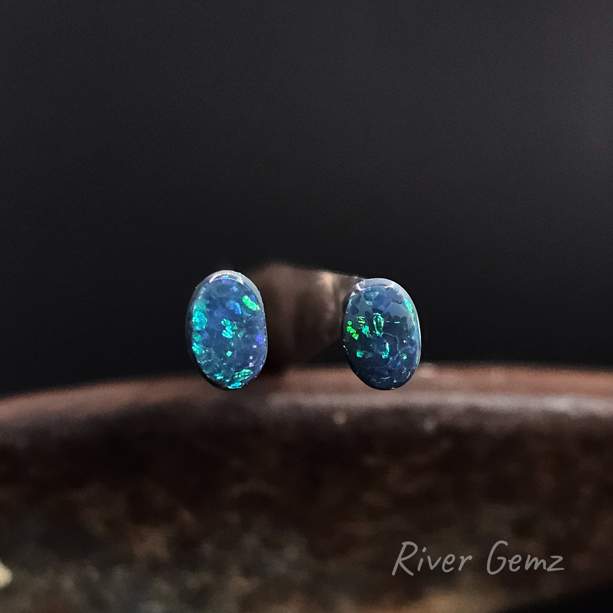A pair of oval unset opals with blue and green colours, displayed on a dark wooden surface with a black background. The brand name 'River Gemz' is visible in the bottom right corner.