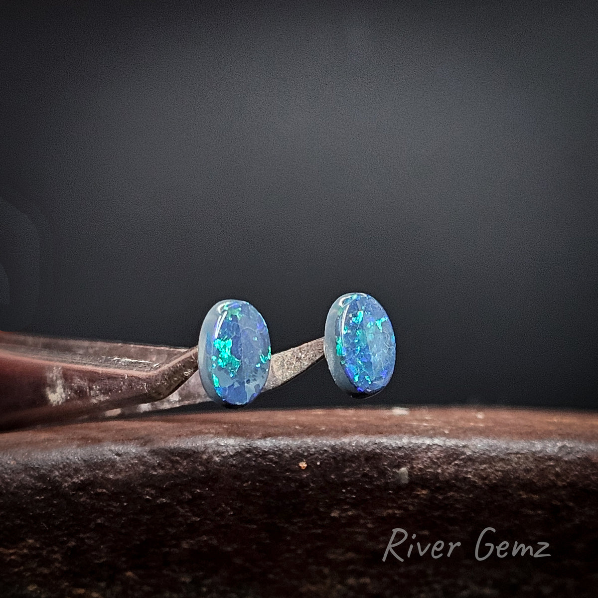 A pair of oval-cut blue opal gemstones displayed on a metal stand against a dark gray background, with a rustic wooden surface below. The brand name 'River Gemz' is visible in the bottom right corner.