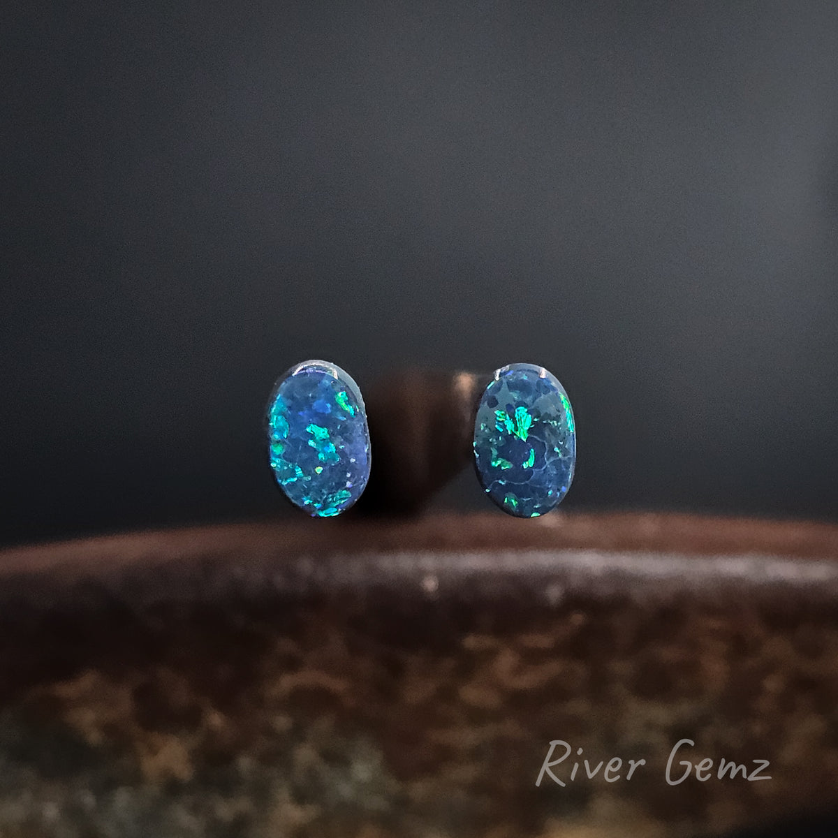 A pair of oval blue and green opals, displayed on a dark brown textured surface against a dark gray background. The watermark 'River Gemz' is visible in the bottom right corner.