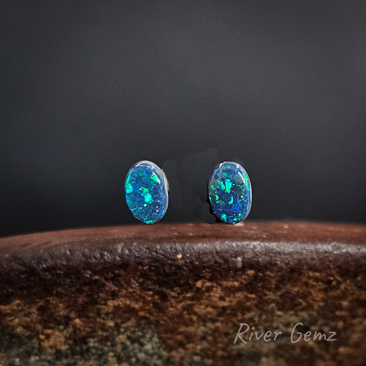 A pair of oval vibrant blue and green opals, resting on a textured brown surface against a dark gray background. The brand name 'River Gemz' is visible as a watermark in the bottom right corner.
