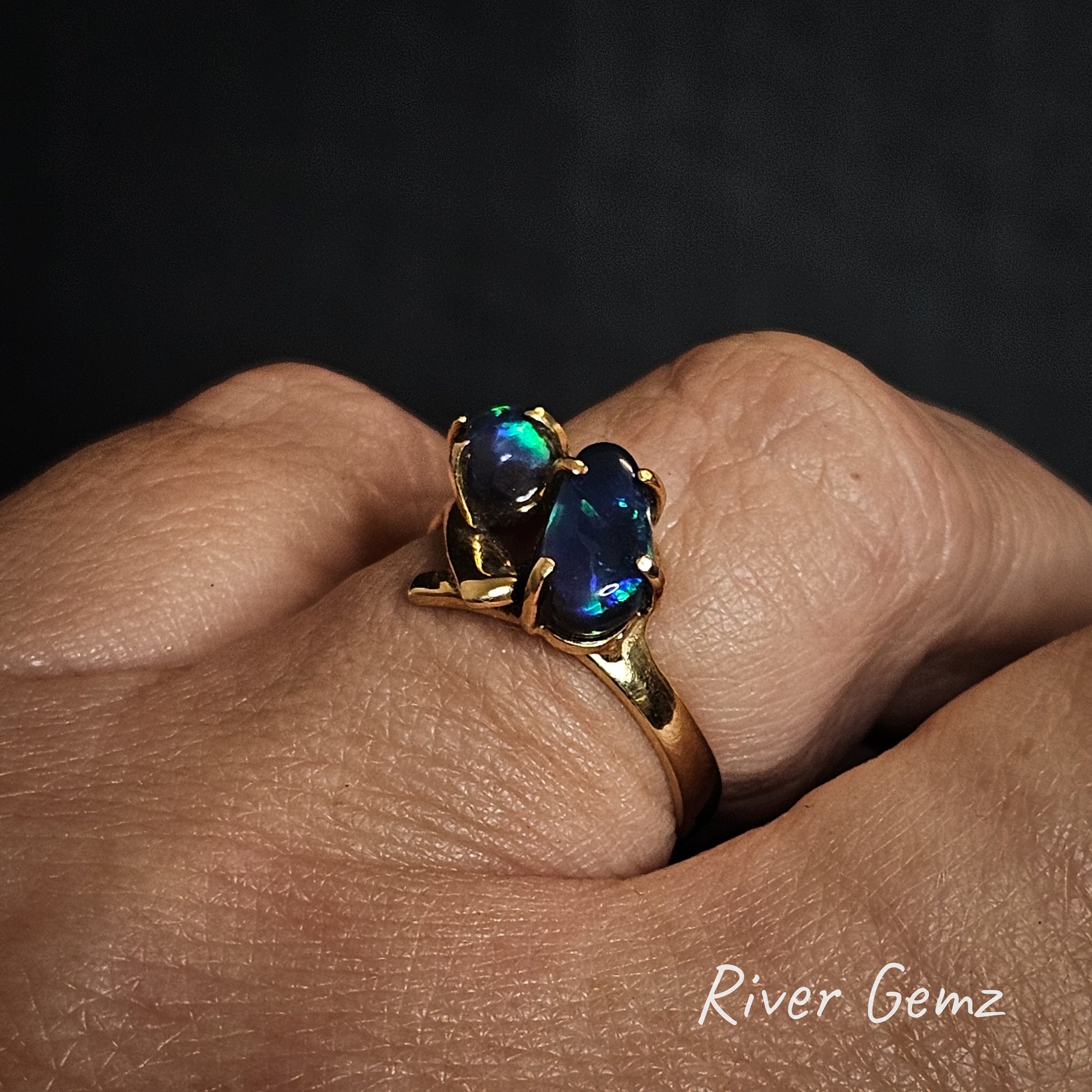 Two dark opals in a gold ring shown worn on a ring finger.