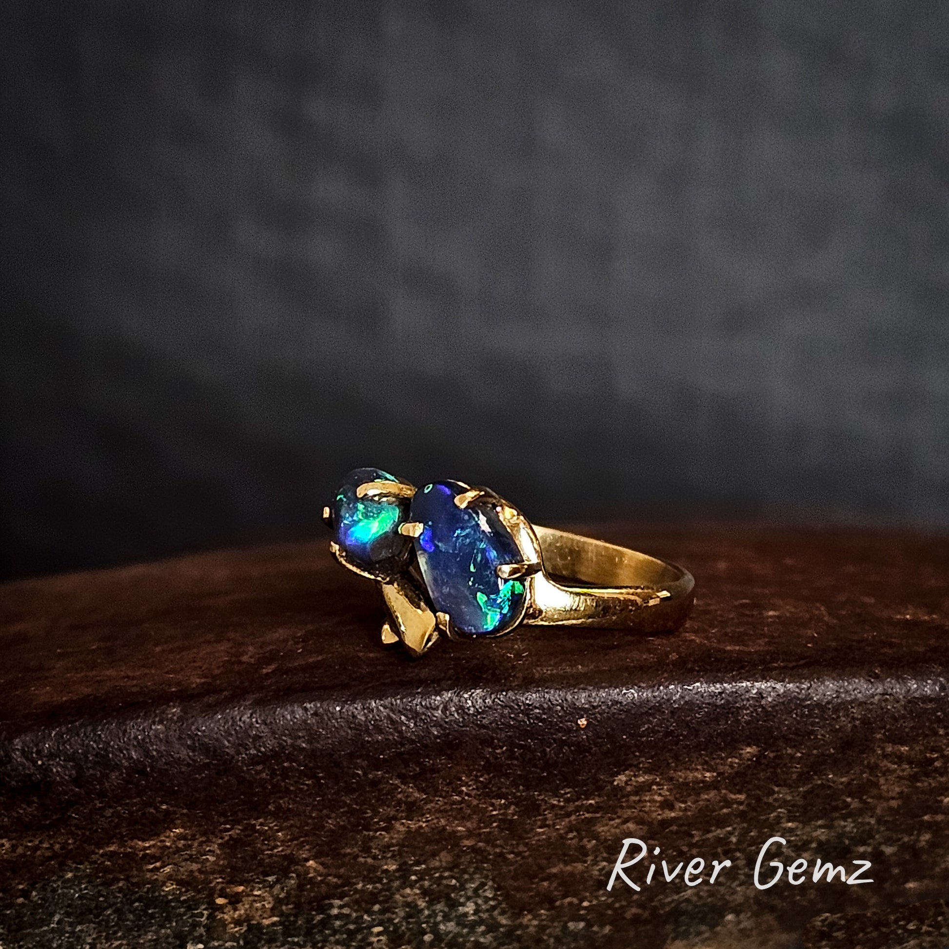 Two dark opals claw set in yellow gold ring on a cast iron surface branded 'River Gemz'.