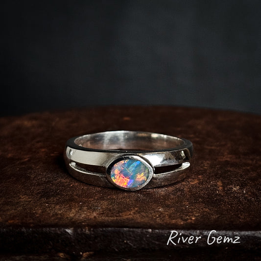 Silver ring with an oval shaped colorful black opal on a dark surface, branded 'River Gemz'.
