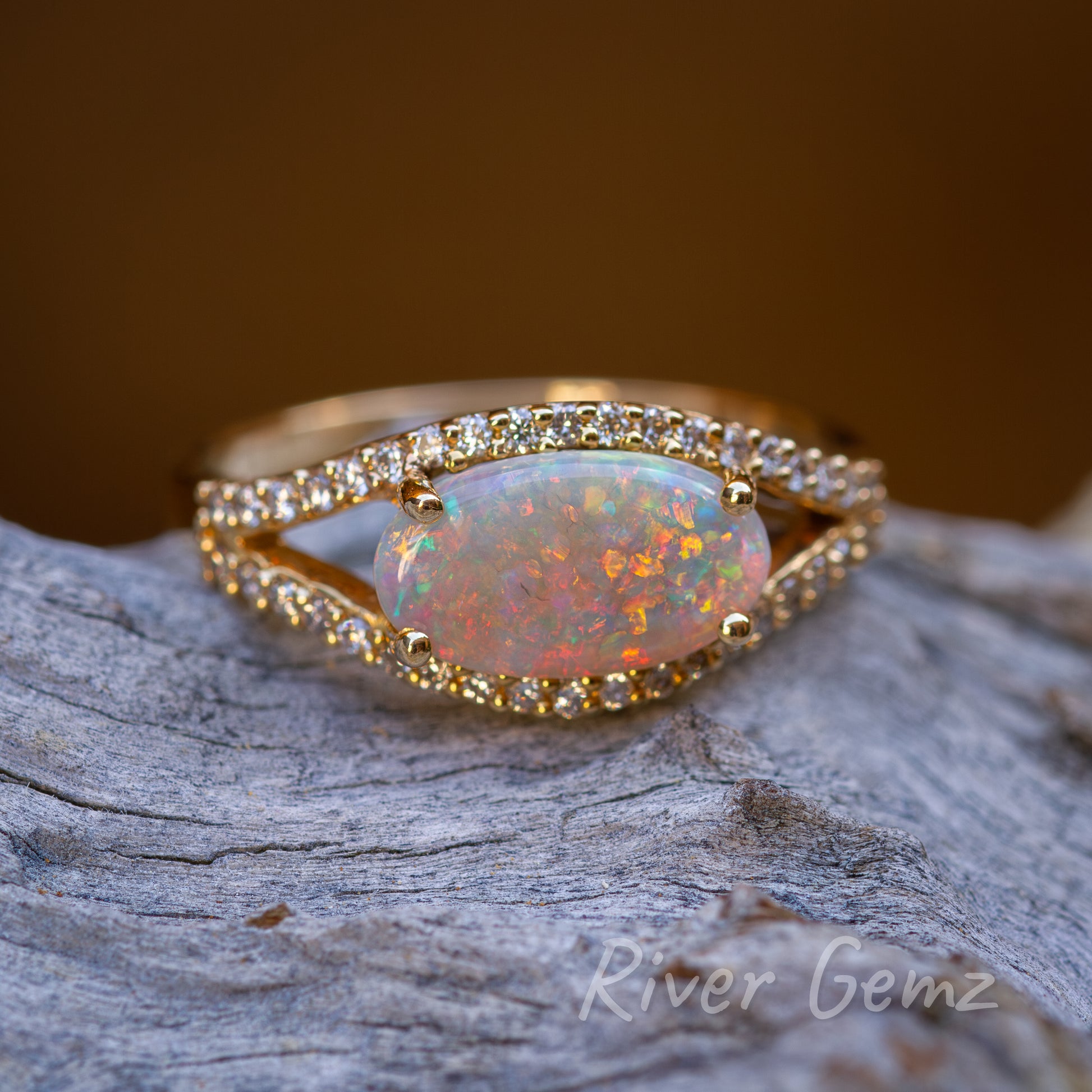 Cabochon multi-coloured crystal opal. The ring design is like an eye with the diamonds forming the shape and the opal the eye ball. The gold ring contrasts well against the weathered light grey piece of driftwood that it is lying on.