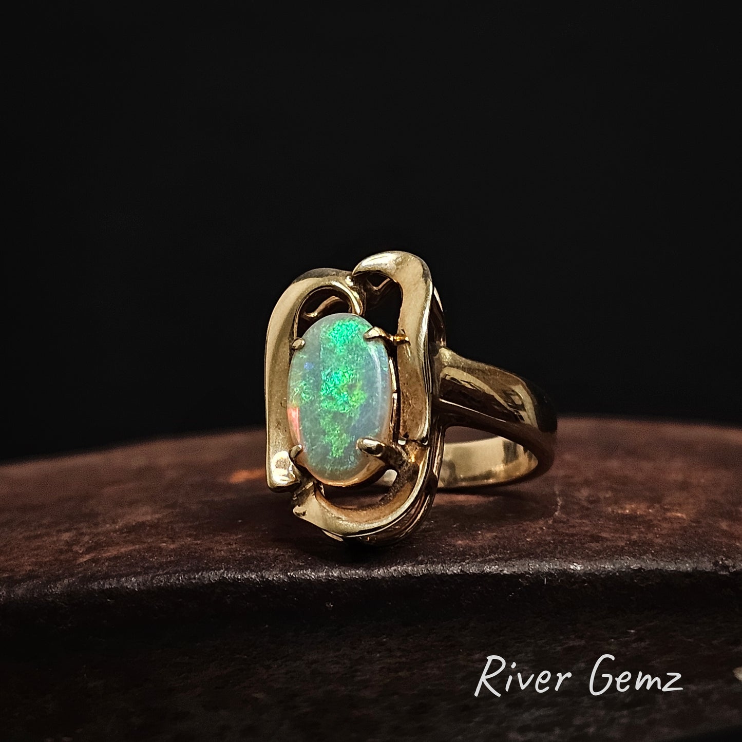 Yellow gold ring with an oval shaped green opal on a dark background, branded 'River Gemz'.