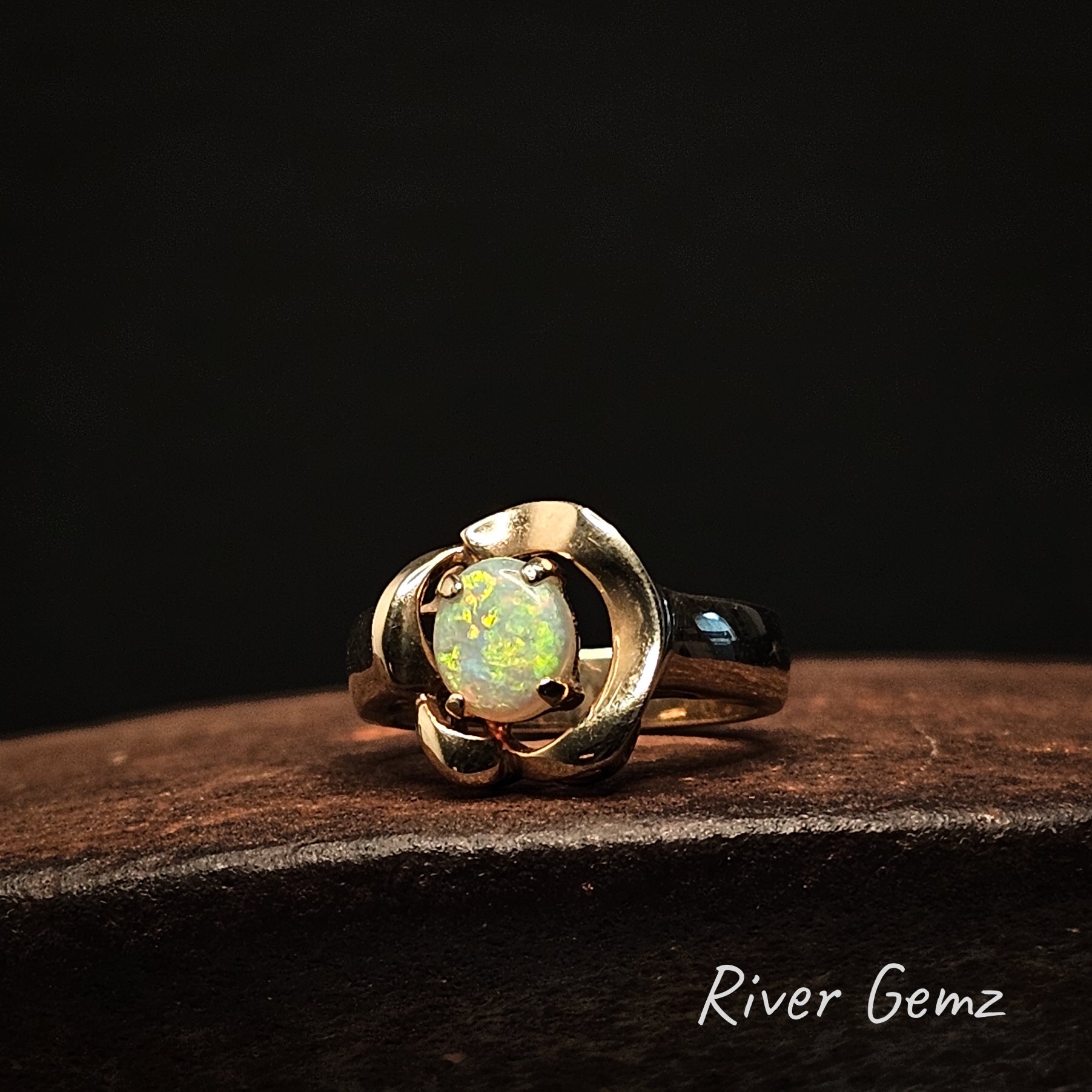 Gold ring with a glowing gemstone on a dark background, featuring 'River Gemz' branding.