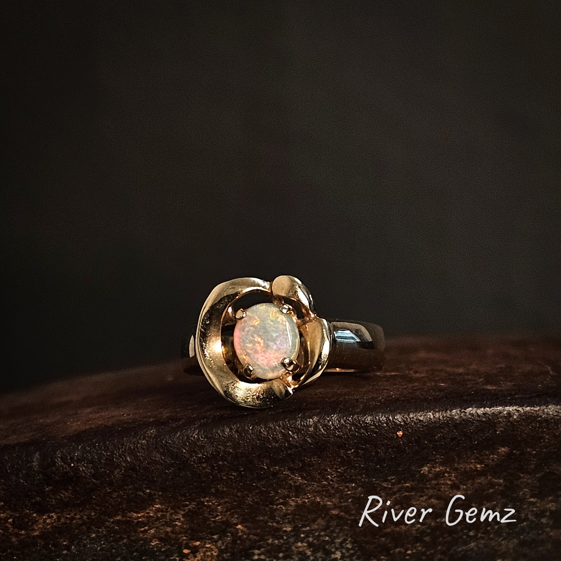 Gold ring with an oval shaped crystal opal on a dark surface, branded 'River Gemz'.