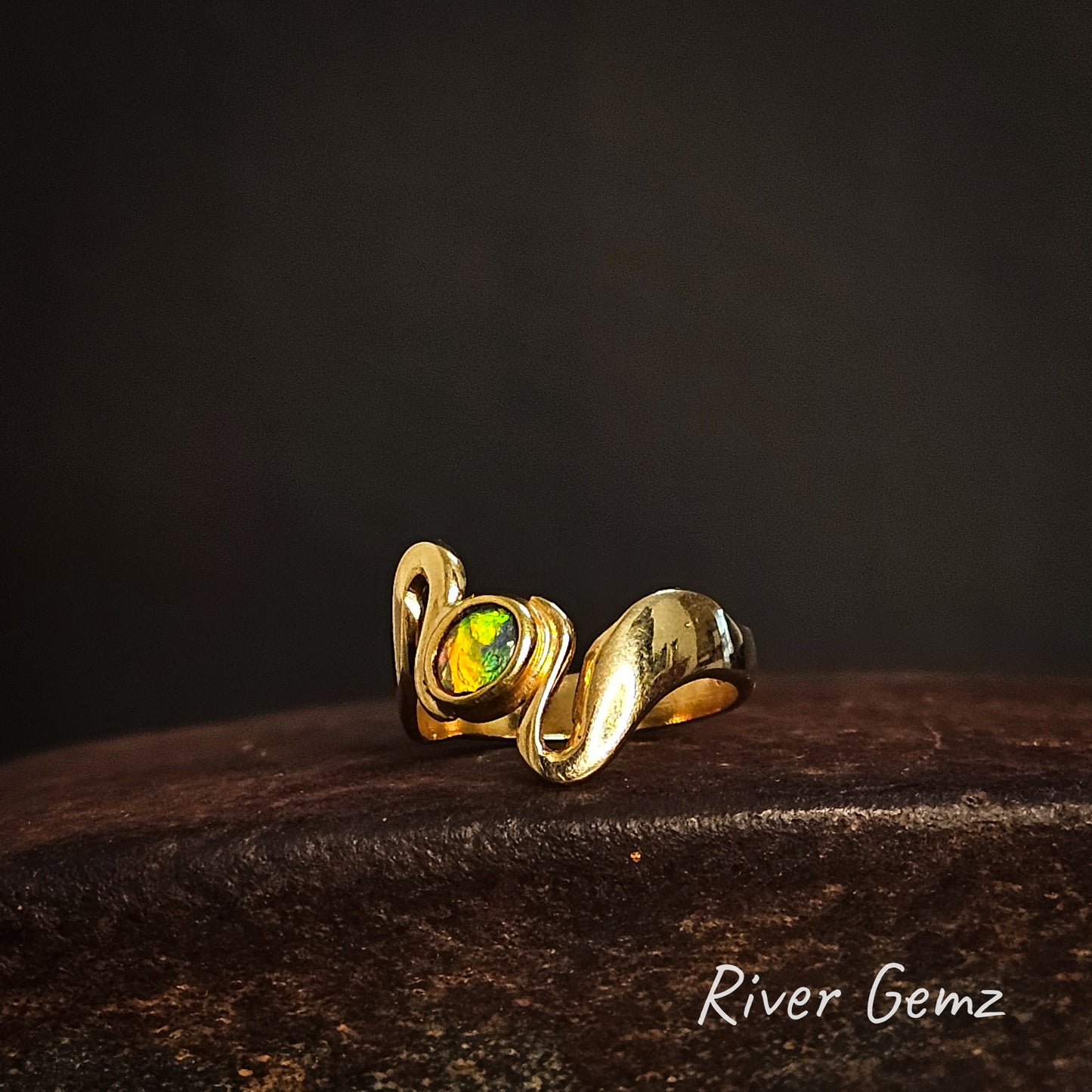 Gold ring with an oval shaped colourful crystal opal on a dark background, featuring the brand 'River Gemz'.