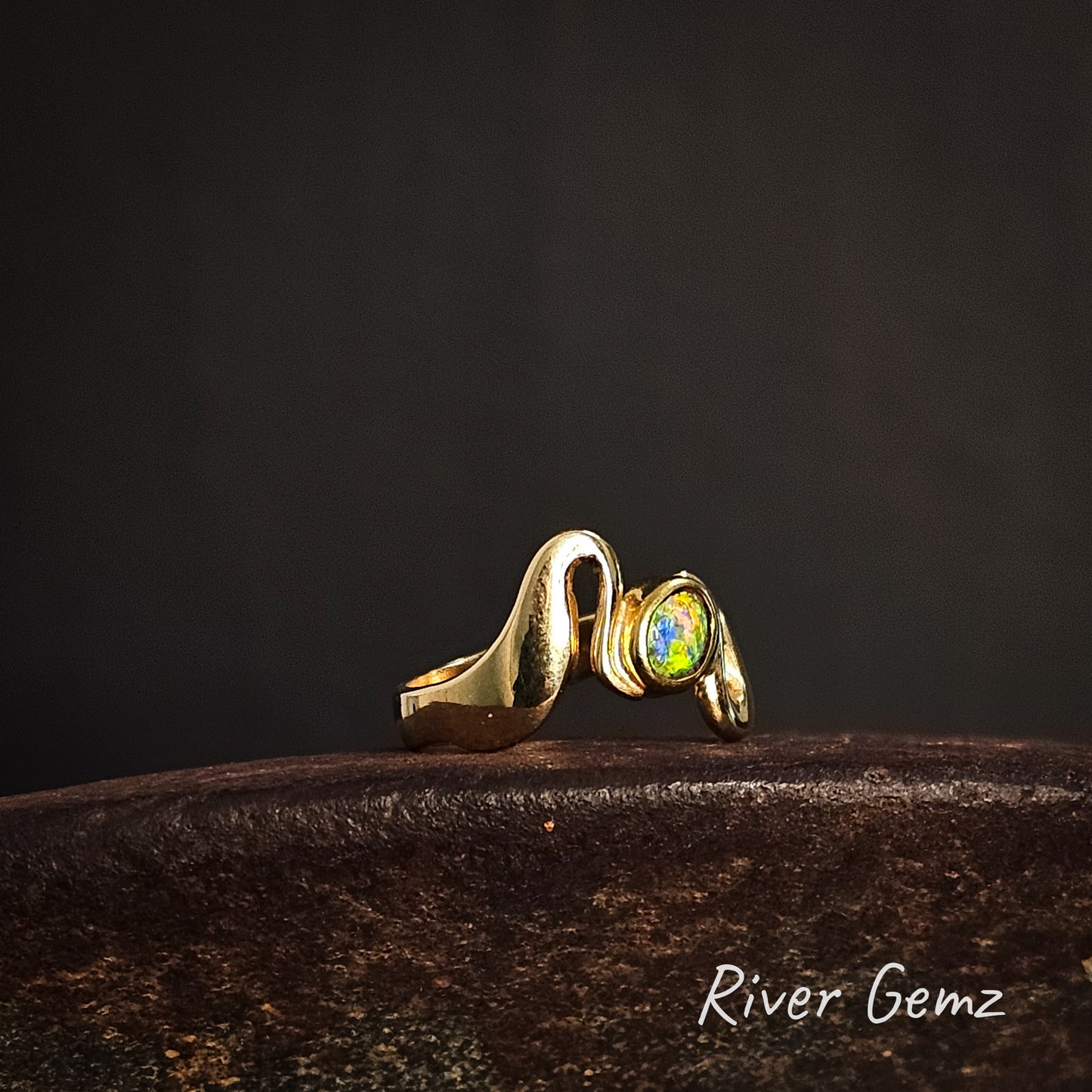 Gold ring with a colorful gemstone on a dark background, featuring the brand 'River Gemz'.